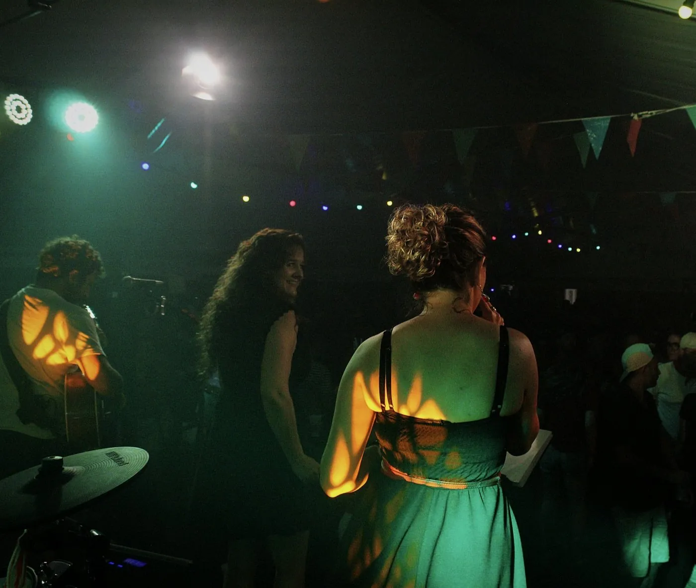 Les deux chanteuses et le guitariste de Watts Up vus de dos sur scène, face au public, sous des fanions et une guirlande lumineuse