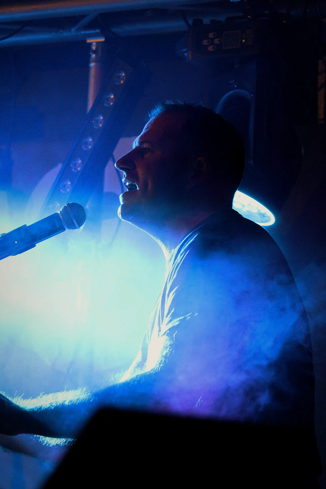 Chanteur de Watts Up de profil au micro, contre-jour bleu et fumée de scène