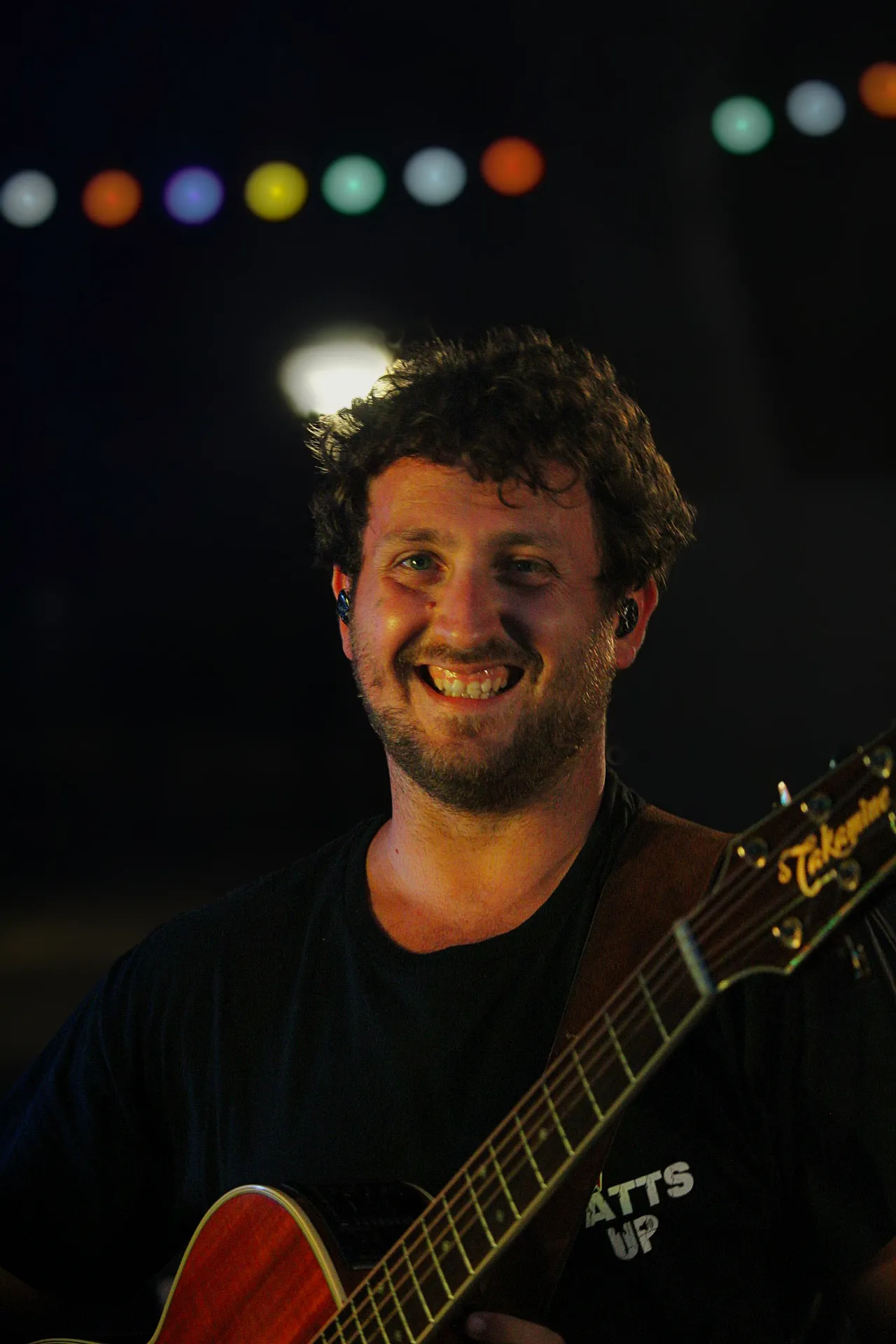 Guitariste de Watts Up tout sourire, t-shirt du groupe, sous les guirlandes des 40 ans de Lous Esquiros